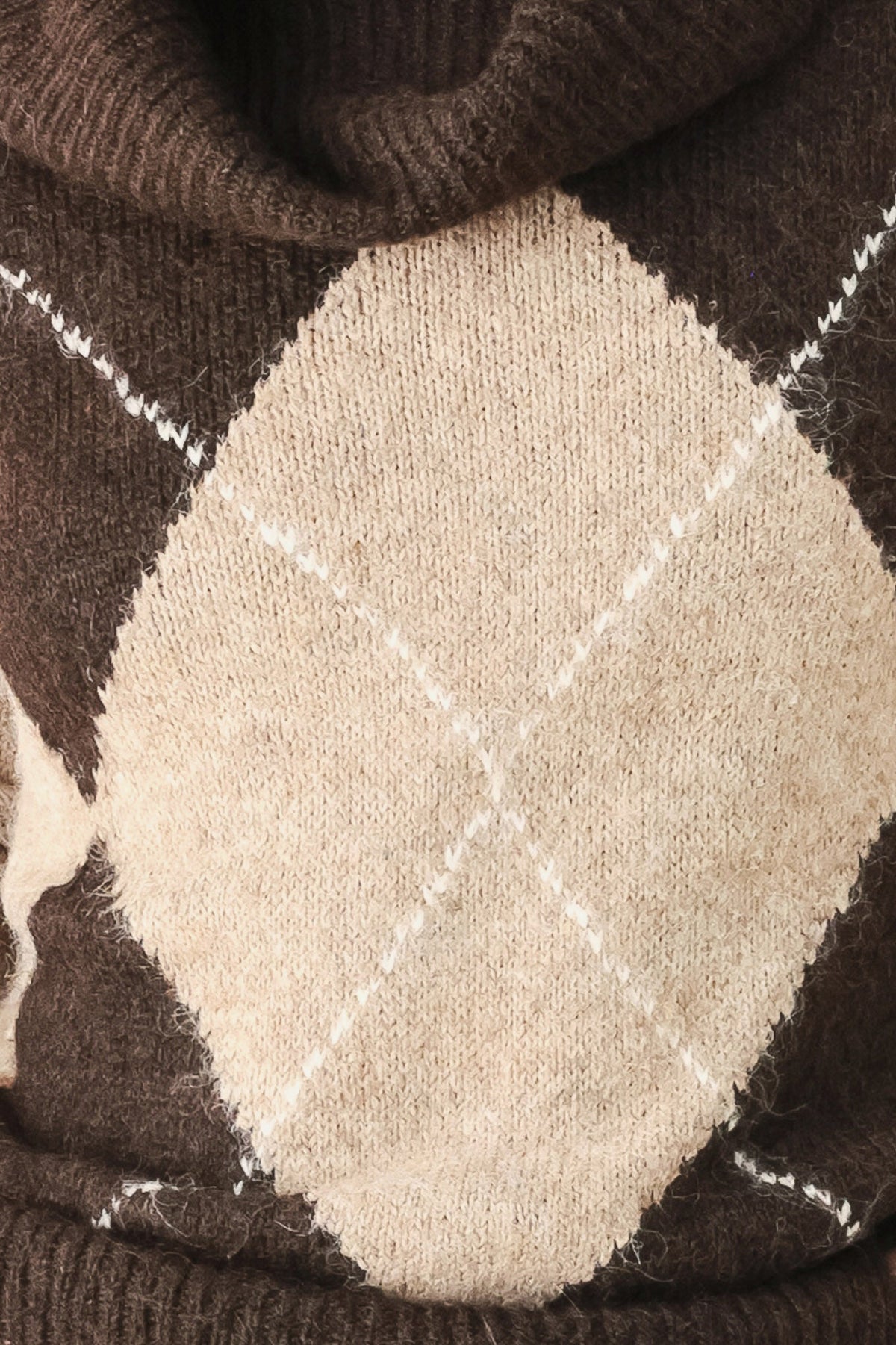 Close-up of a knit sweater showing an argyle pattern with a large beige diamond, dark brown sections, and thin white diagonal stripes.