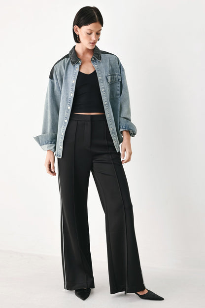 A model wearing a light blue denim button-up shirt with black shoulder patches, worn open over a black crop top and black wide-leg trousers with white side stripes, set against a plain white studio background.