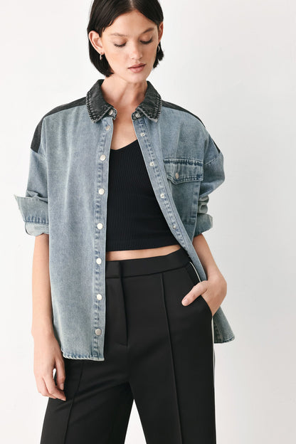 A model wearing a light-wash blue denim button-up shirt with dark gray contrast panels on the shoulders and collar, styled over a black ribbed tank top and black trousers, against a plain light gray background.