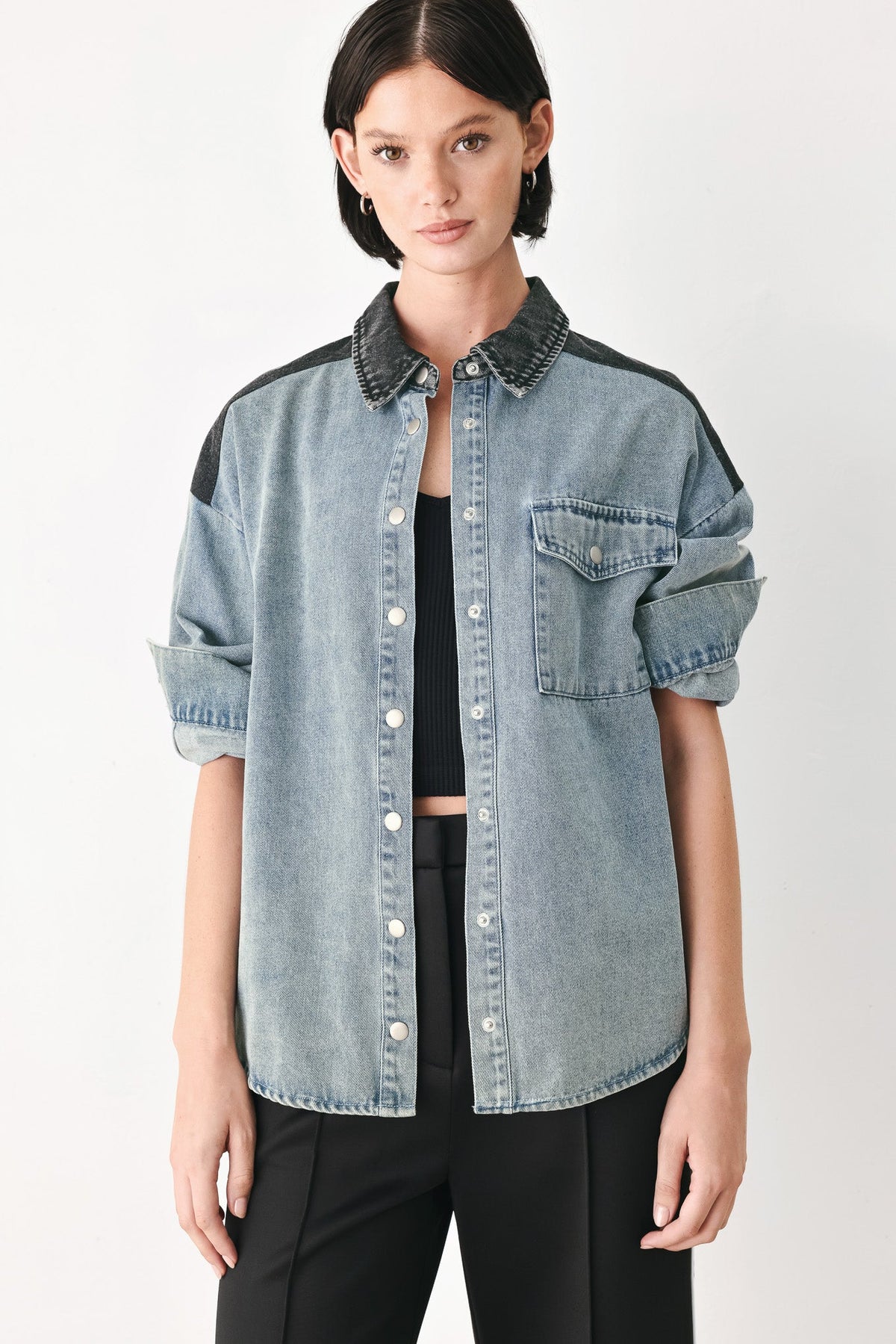 A model wearing a light-wash denim button-up shirt with dark charcoal gray contrast panels on the shoulders and collar, styled over a black crop top and black trousers against a light gray studio background.