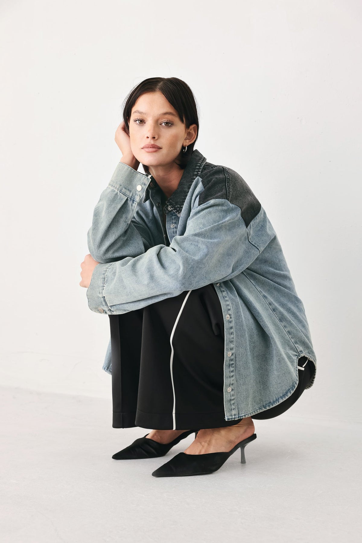 A model crouches in a studio setting wearing a light blue oversized denim jacket over a black dress and black pointed-toe heels.