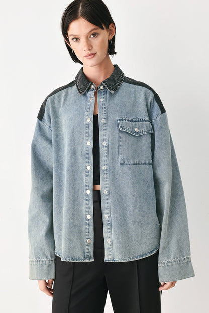 A model wearing a light-wash denim button-up shirt jacket with black contrast panels on the shoulders and collar, styled over a black top and black trousers against a light gray studio background.