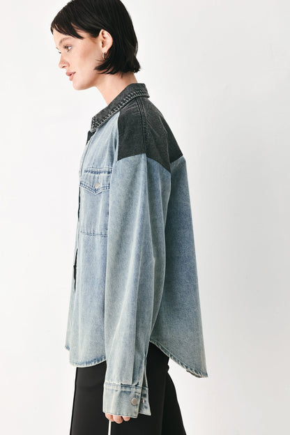 A model shown in profile wearing a light blue denim shirt jacket with dark charcoal gray color-blocked shoulder panels, styled with black pants against a plain white studio background.