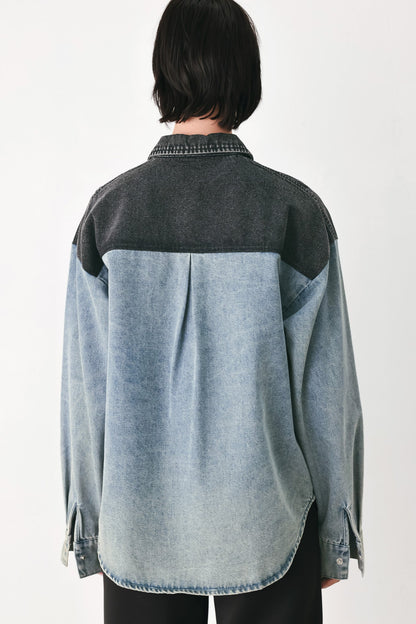 A person shown from the back wearing a long-sleeve denim shirt with a dark charcoal gray yoke and light blue washed lower body, paired with black pants against a plain light background.