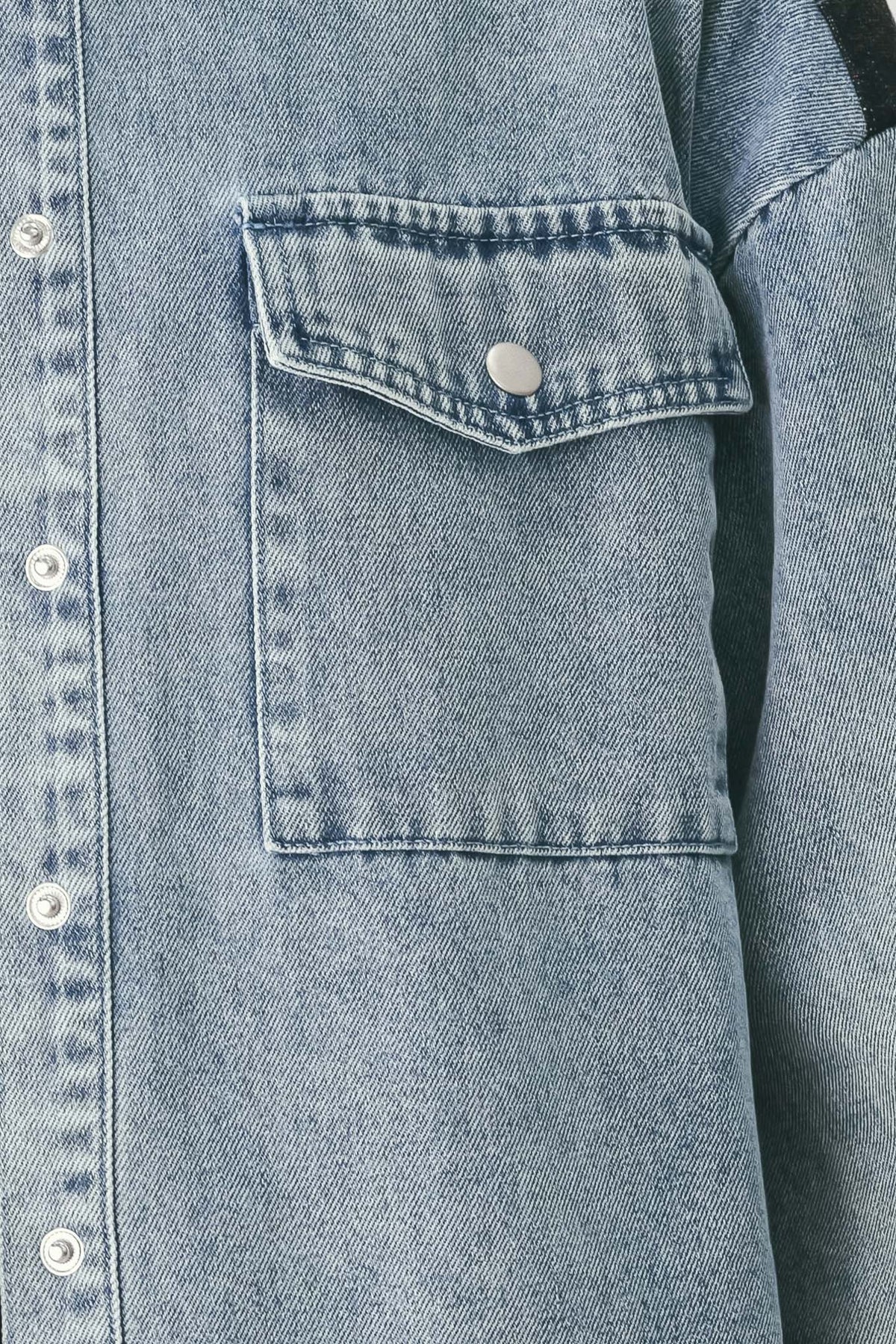Close-up of a light blue denim jacket showing a buttoned chest pocket with a silver snap button and white buttons on the front placket.
