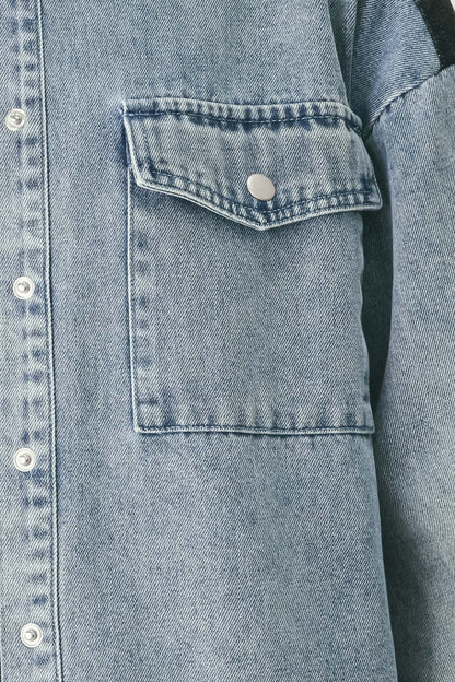 Close-up of a light blue denim jacket showing a buttoned chest pocket with a silver snap button and white buttons on the front placket.