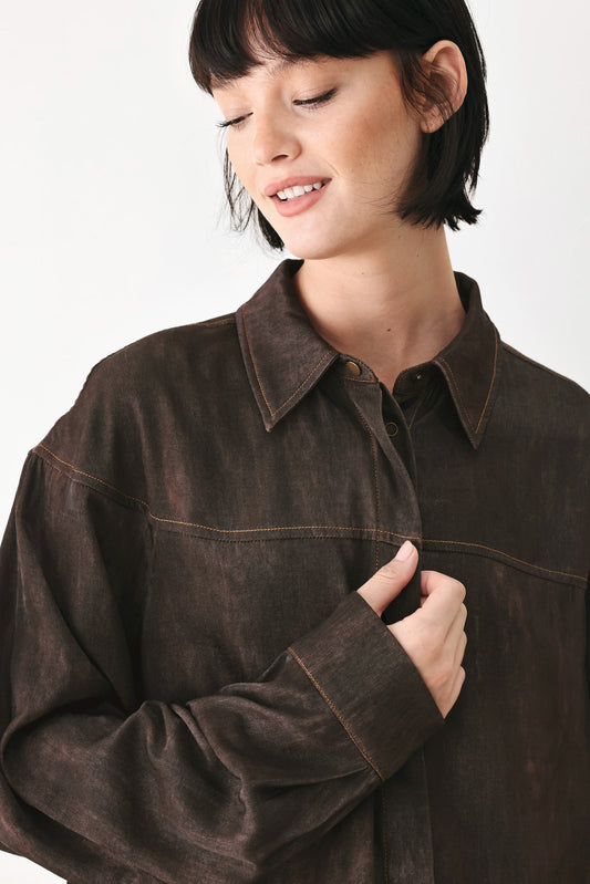 A woman wearing a dark brown, washed-finish button-up shirt jacket with contrast stitching, shown from the chest up against a plain white background.