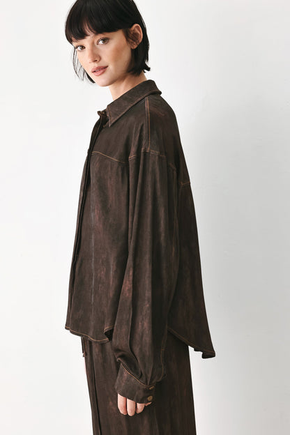 A model wearing a dark brown, long-sleeved, oversized button-up shirt with a distressed, washed texture, paired with matching dark brown trousers, set against a plain light gray background.