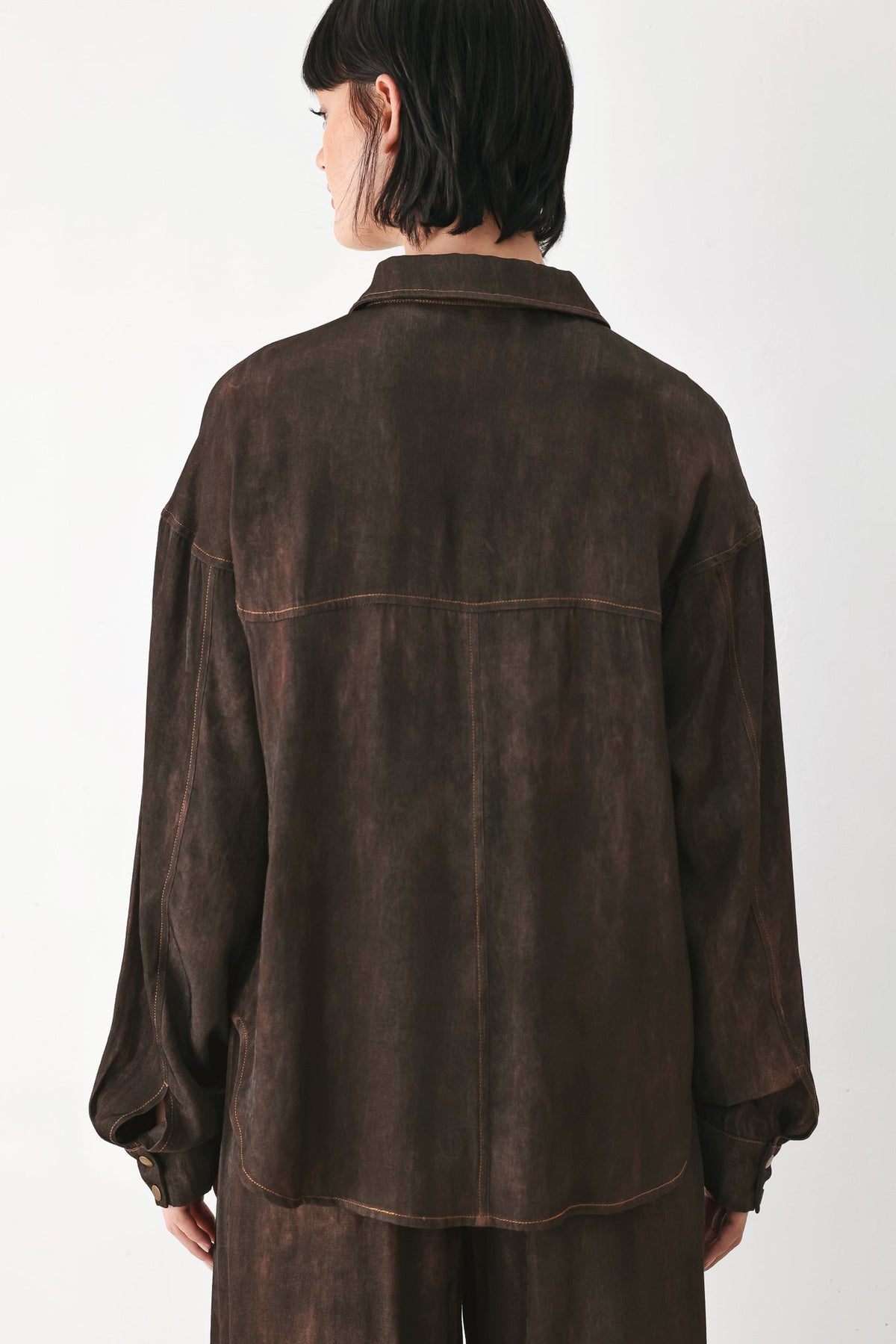 A model viewed from the back wearing a dark brown, washed-texture button-up shirt jacket and matching trousers against a plain white studio background.