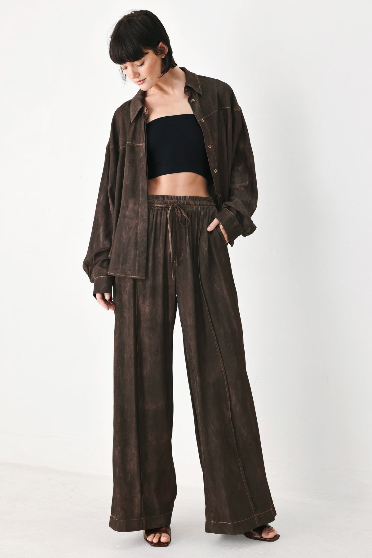 A model wearing a dark brown washed denim outfit consisting of a long-sleeve button-up shirt and matching wide-leg drawstring pants, styled over a black bandeau top and brown sandals, against a plain white studio background.