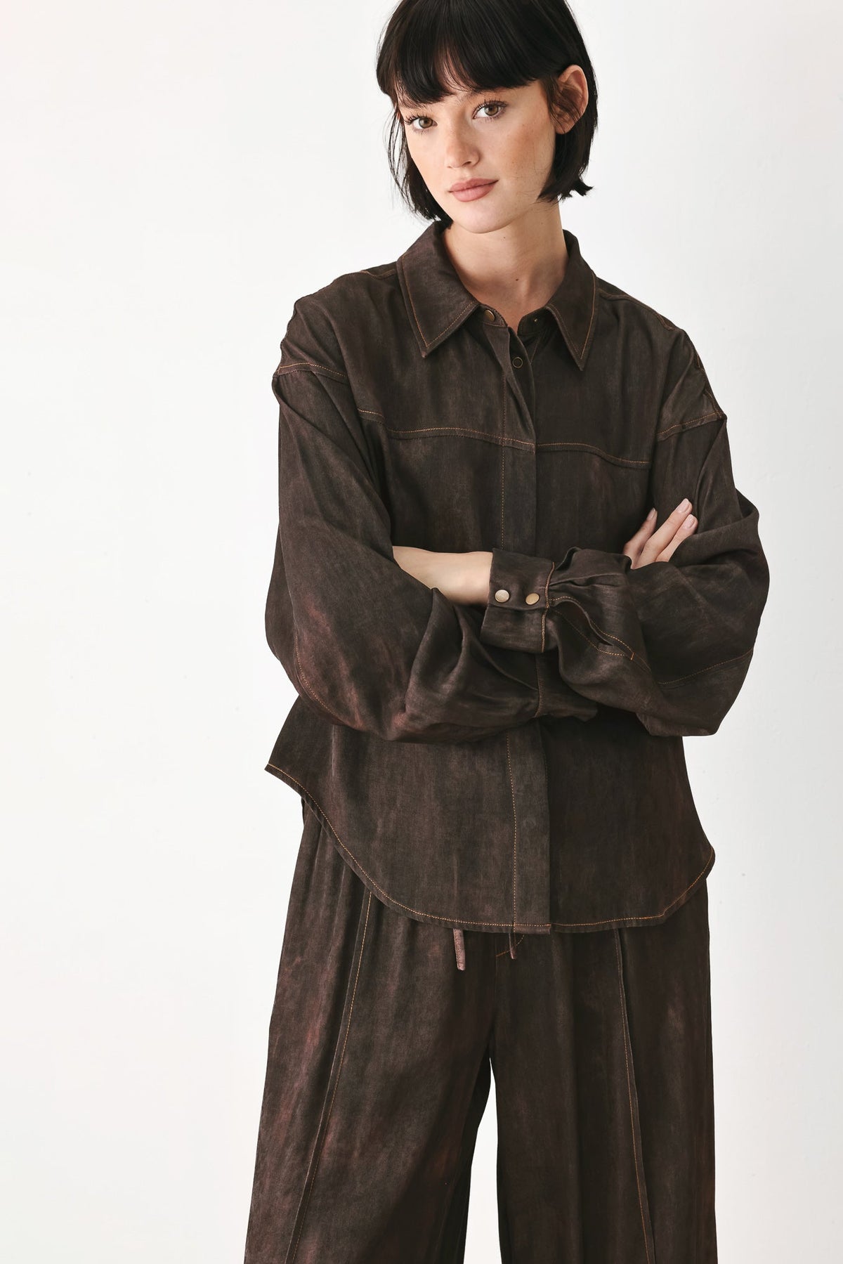 A model wearing a dark brown washed cotton shirt jacket and matching wide-leg trousers, standing with arms crossed against a plain white studio background.
