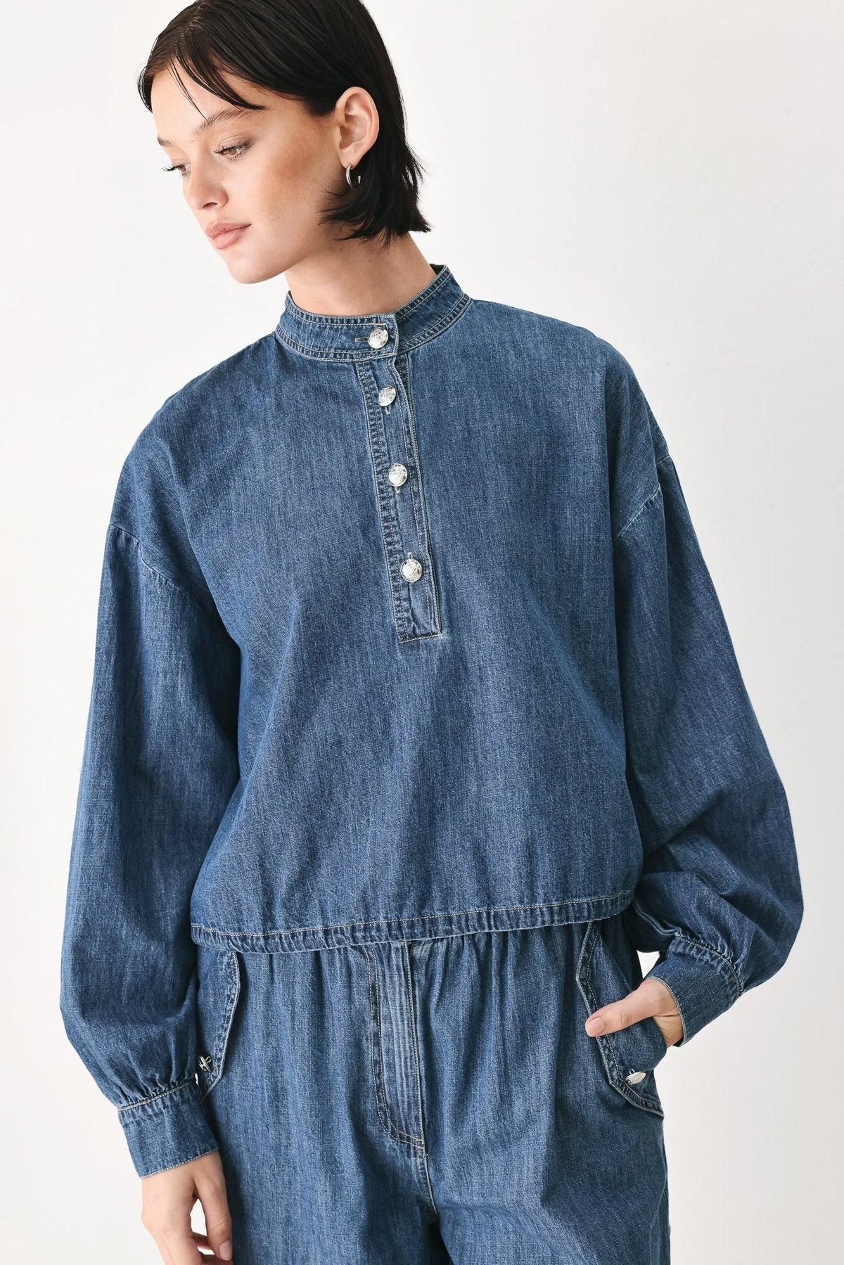 A model wearing a blue denim long-sleeve jumpsuit with a buttoned stand collar and elastic waist, photographed against a plain light background.