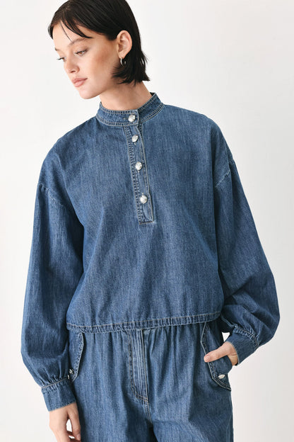 A model wearing a blue denim long-sleeve jumpsuit with a buttoned stand collar and elastic waist, photographed against a plain light background.