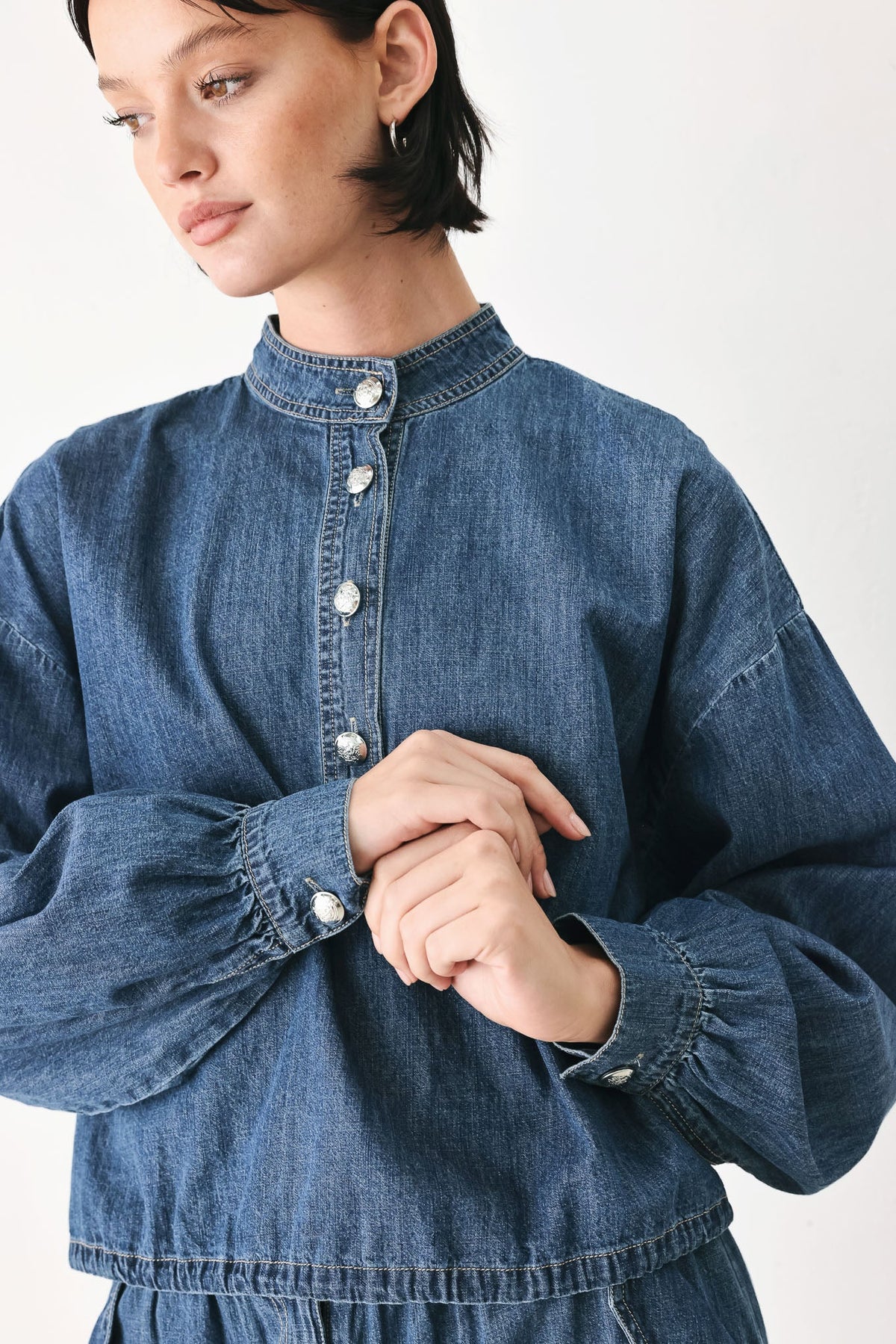 A close-up of a model wearing a blue denim long-sleeve shirt with a high collar and decorative silver buttons down the front and on the cuffs.