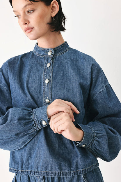 A close-up of a model wearing a blue denim long-sleeve shirt with a high collar and decorative silver buttons down the front and on the cuffs.