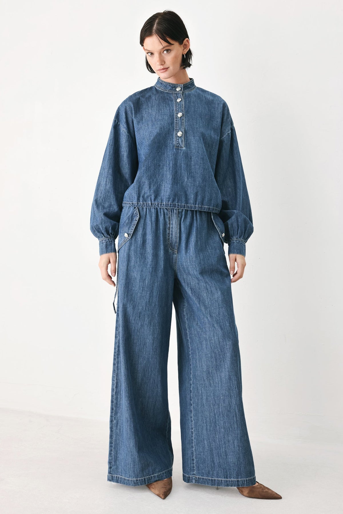 A model wearing a blue denim one-piece jumpsuit with a high collar, buttoned chest, and wide-leg pants, standing against a plain white studio background.