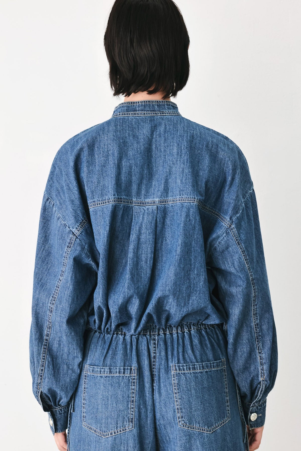 A person viewed from the back wearing a blue denim jumpsuit with long sleeves, a buttoned collar, and two large patch pockets at the back, against a plain light background.