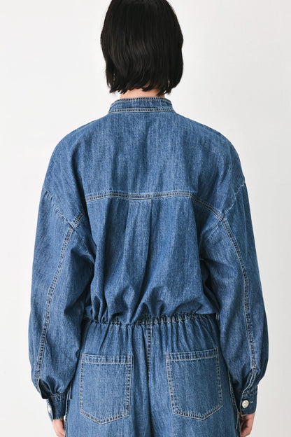 A person viewed from the back wearing a blue denim jumpsuit with long sleeves, a buttoned collar, and two large patch pockets at the back, against a plain light background.