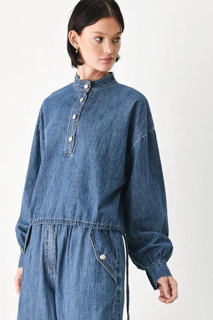 A model wearing a blue denim jumpsuit with a high collar, buttoned placket, and voluminous long sleeves, photographed against a plain white studio background.