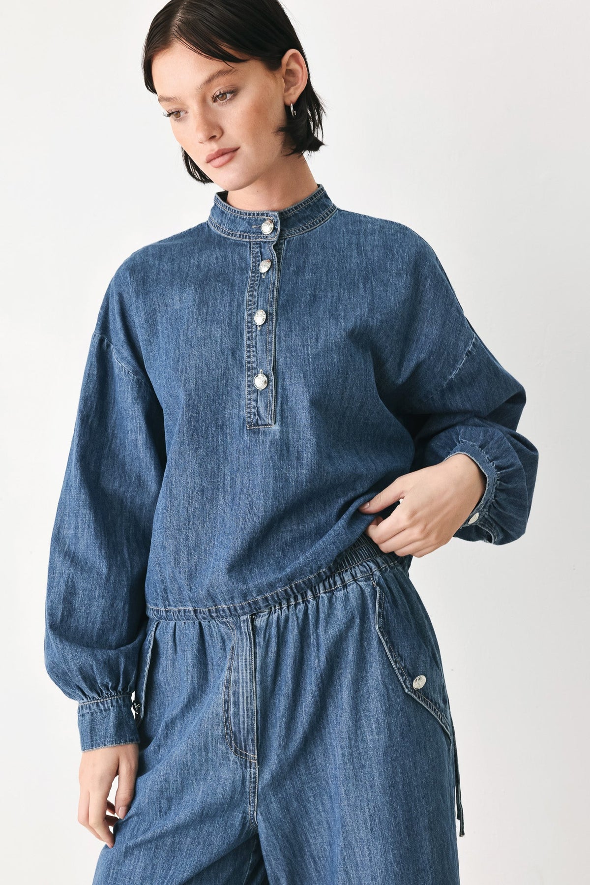 A model wearing a blue denim jumpsuit with a high mock neck, buttoned placket, and elastic waist, photographed against a plain light gray studio background.