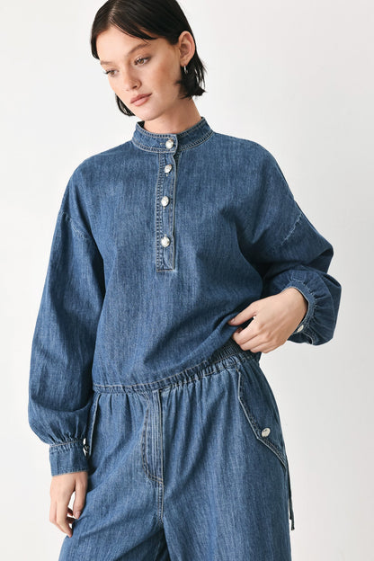 A model wearing a blue denim jumpsuit with a high mock neck, buttoned placket, and elastic waist, photographed against a plain light gray studio background.