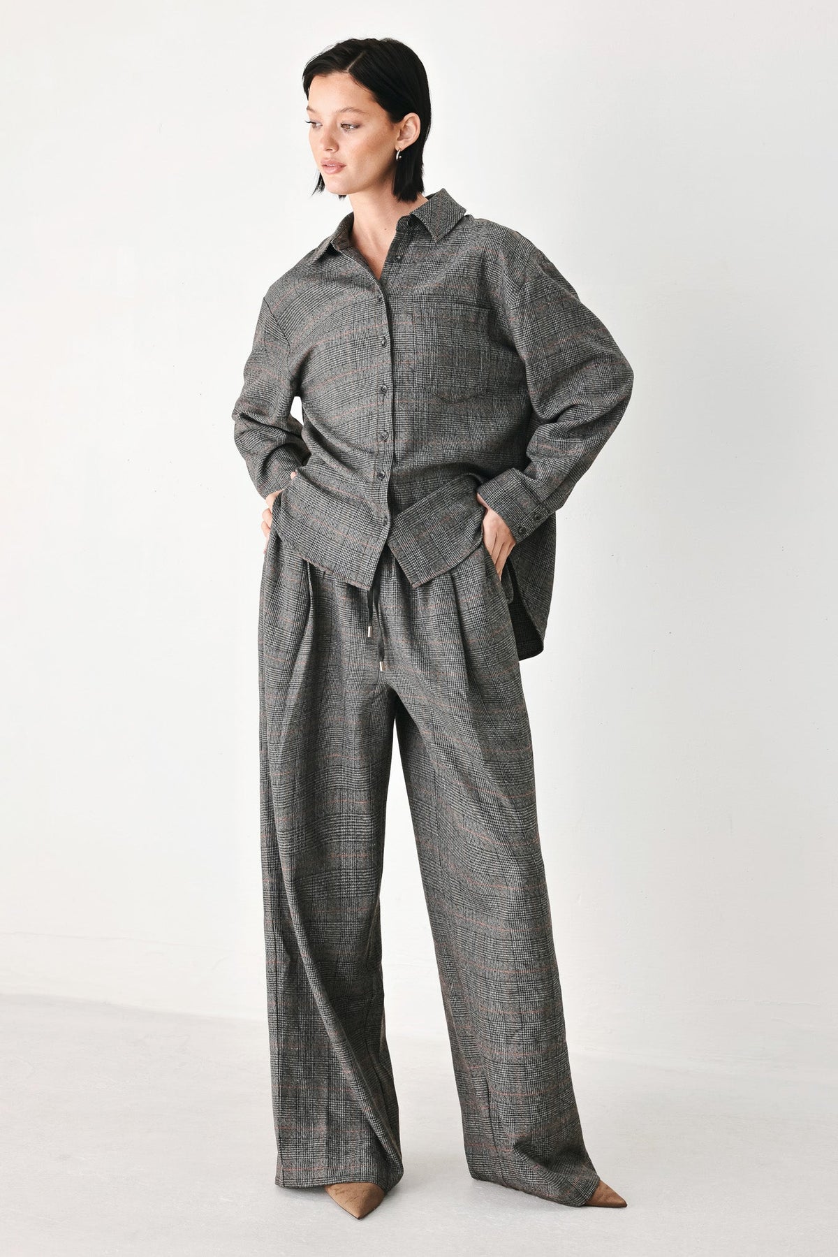 A model wearing a matching gray plaid button-up shirt and wide-leg trousers set, standing against a plain light gray studio background.