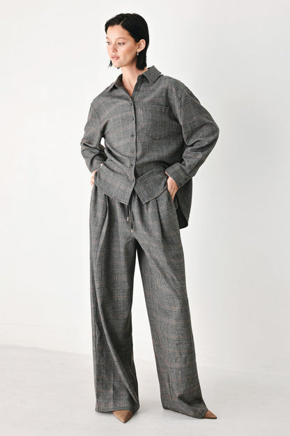 A model wearing a matching gray plaid button-up shirt and wide-leg trousers set, standing against a plain light gray studio background.