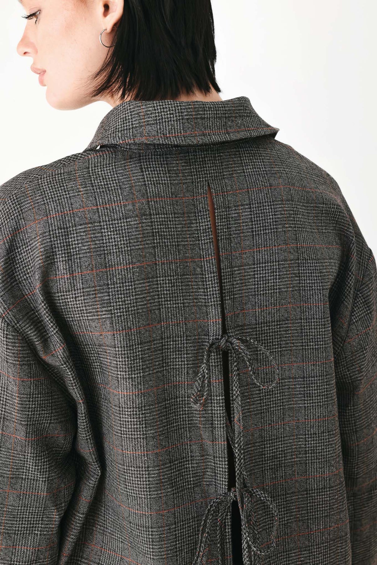 Close-up back view of a person wearing a dark gray plaid overshirt with a tie closure at the back, against a plain light background.