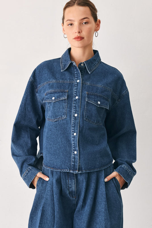 A woman wearing a dark blue denim button-up shirt jacket with two chest pockets, paired with matching denim bottoms, against a plain light gray studio background.