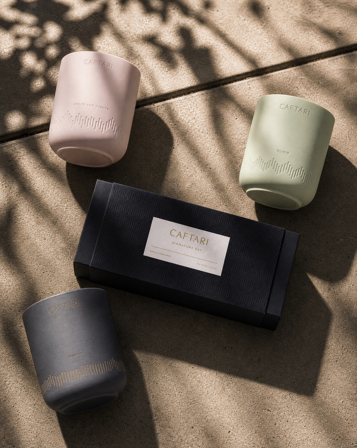 Three Caftari candles in pink, sage green and charcoal around a black gift box on a sunlit stone surface