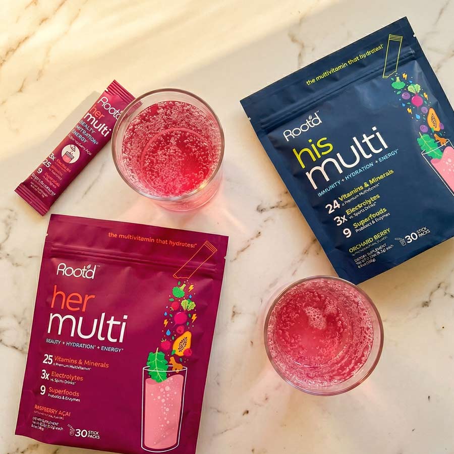 Two packets of Rootd 'her multi' and 'his multi' with two glasses of pink drink on a marble surface.