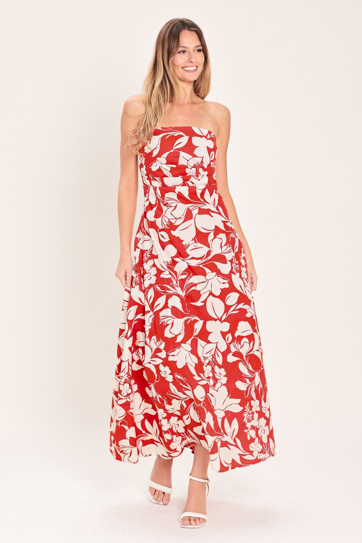 A woman modeling a strapless red maxi dress with a white tropical floral print, paired with white heeled sandals, against a solid off-white studio background.