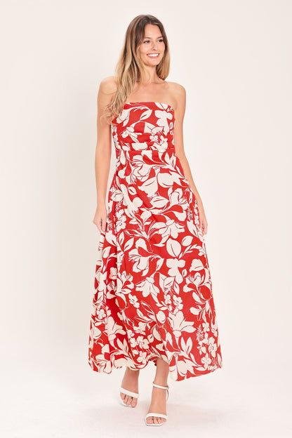 A woman modeling a strapless red maxi dress with a white tropical floral print, paired with white heeled sandals, against a solid off-white studio background.