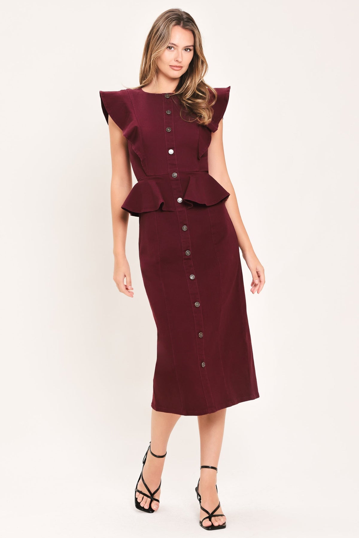 A woman modeling a sleeveless midi dress in deep burgundy. The dress features a button-down front, a peplum waist, and ruffled cap sleeves, paired with black strappy high-heeled sandals against a plain off-white studio background.