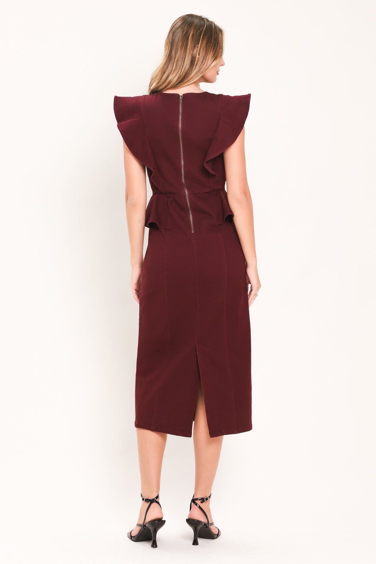 A woman viewed from the back wearing a sleeveless burgundy midi dress with dramatic ruffled shoulders, a front slit, and a central zipper. She is wearing black strappy heels against a plain white studio background.