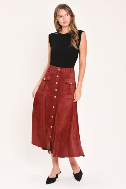 A woman modeling a sleeveless black top and a high-waisted rust-brown midi skirt made of a suede-like material with gold-tone buttons down the front and flap pockets, paired with black pointed-toe heels, against a plain white studio background.