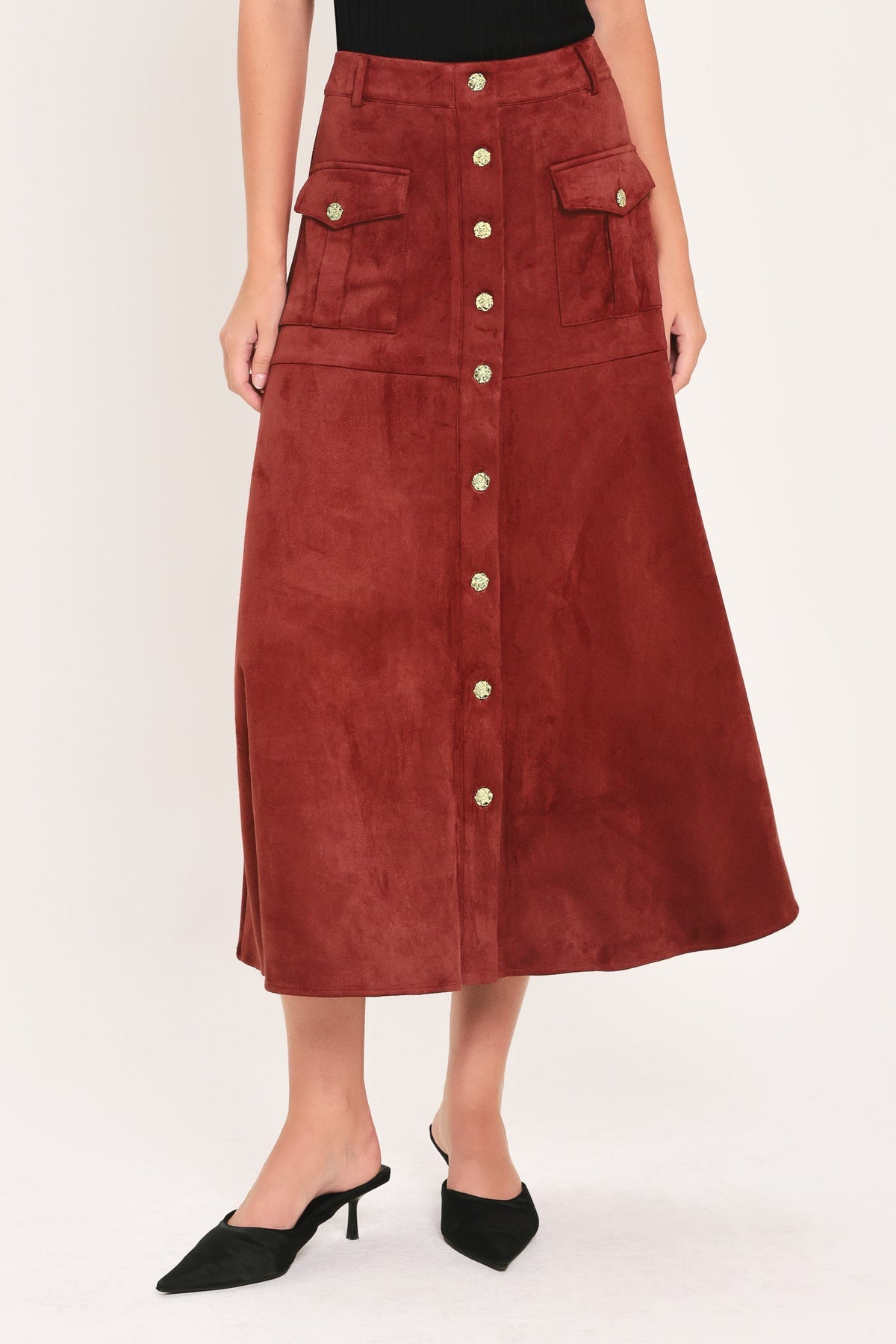 A rust-brown suede midi skirt with a button-down front, flap pockets, and gold-tone button accents, shown on a model wearing black pointed-toe mule heels against a light studio background.