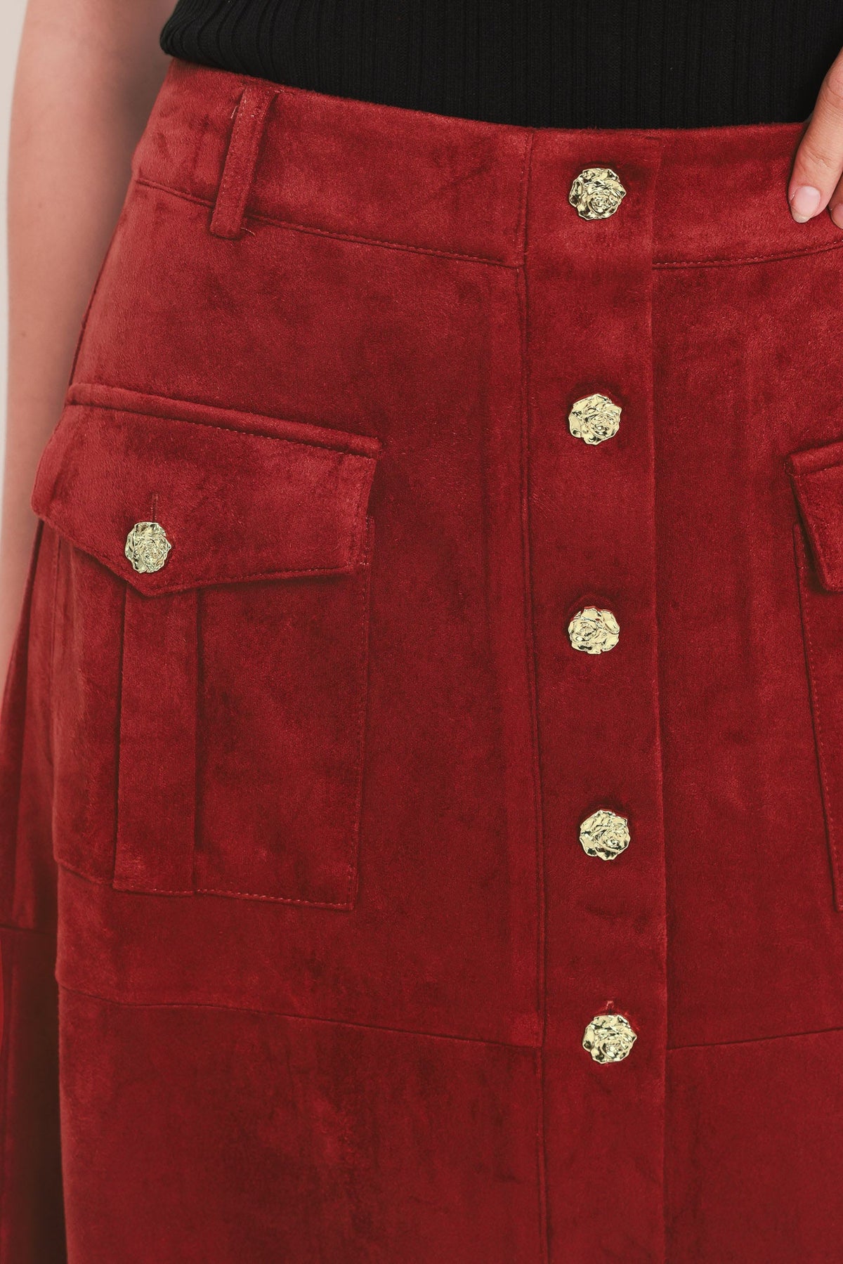 A close-up of a red suede skirt featuring a button-front closure with ornate gold buttons and two side flap pockets with matching gold buttons, worn with a black top.