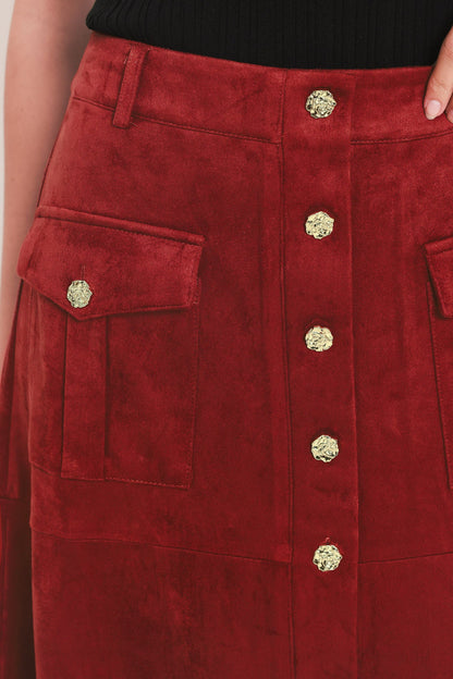 A close-up of a red suede skirt featuring a button-front closure with ornate gold buttons and two side flap pockets with matching gold buttons, worn with a black top.