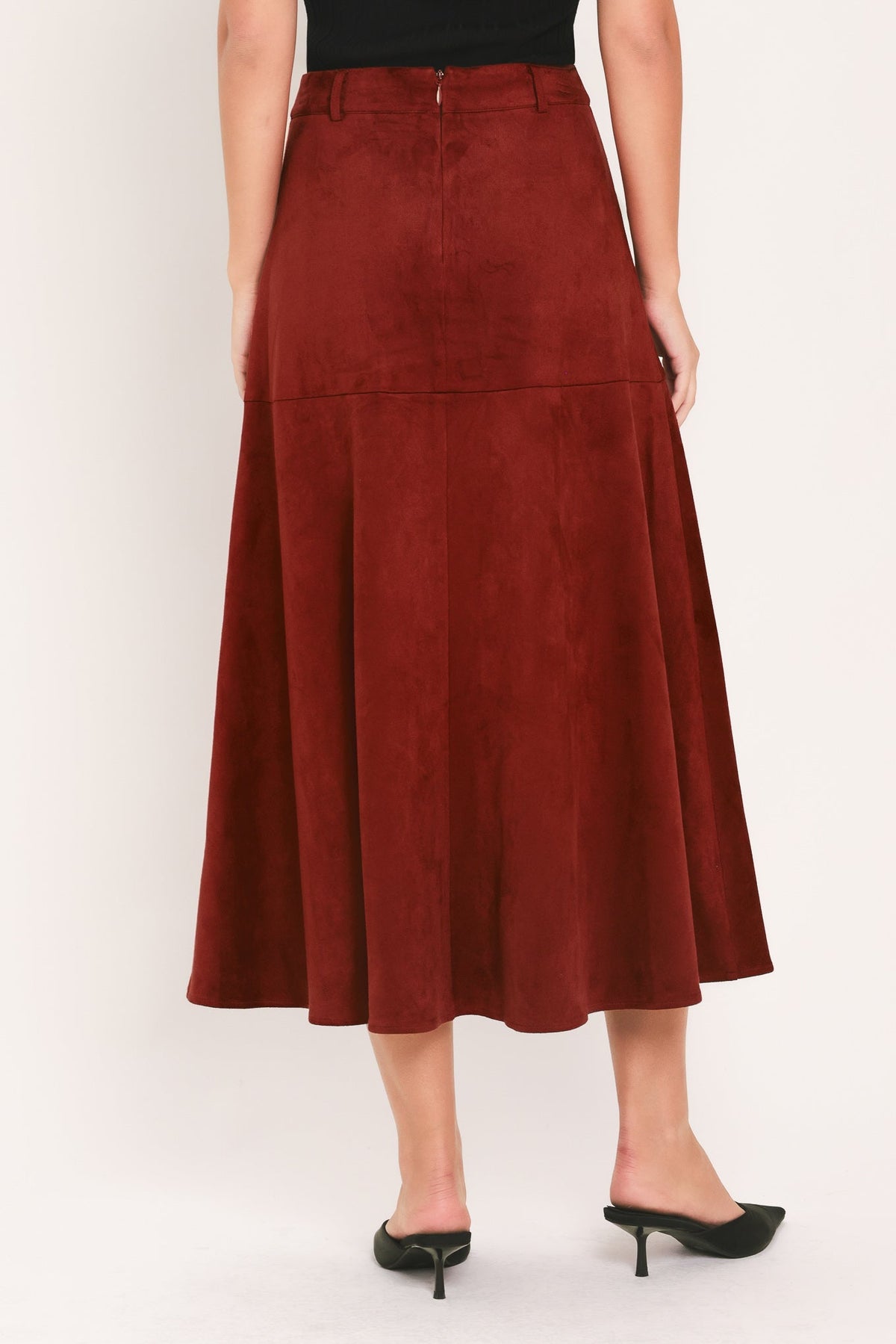 A woman wearing a rust-brown suede midi skirt with a black top and black pointed-toe mule heels, shown from the waist down against a plain off-white studio background.