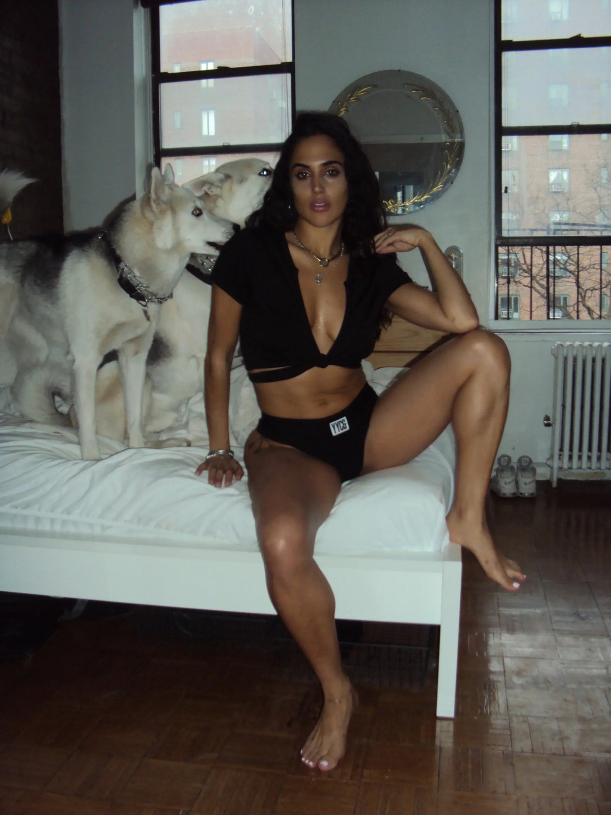 Woman in black lingerie sitting on a bed with a dog beside her in a room with large windows.