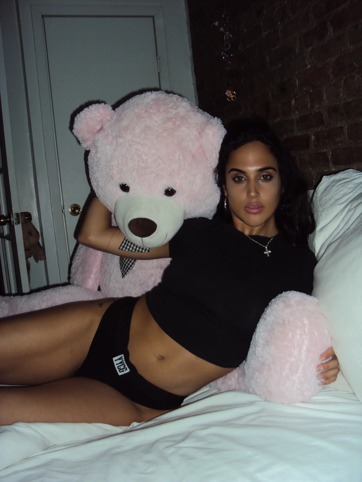 Person in black top and underwear holding a large pink teddy bear in a bedroom setting.