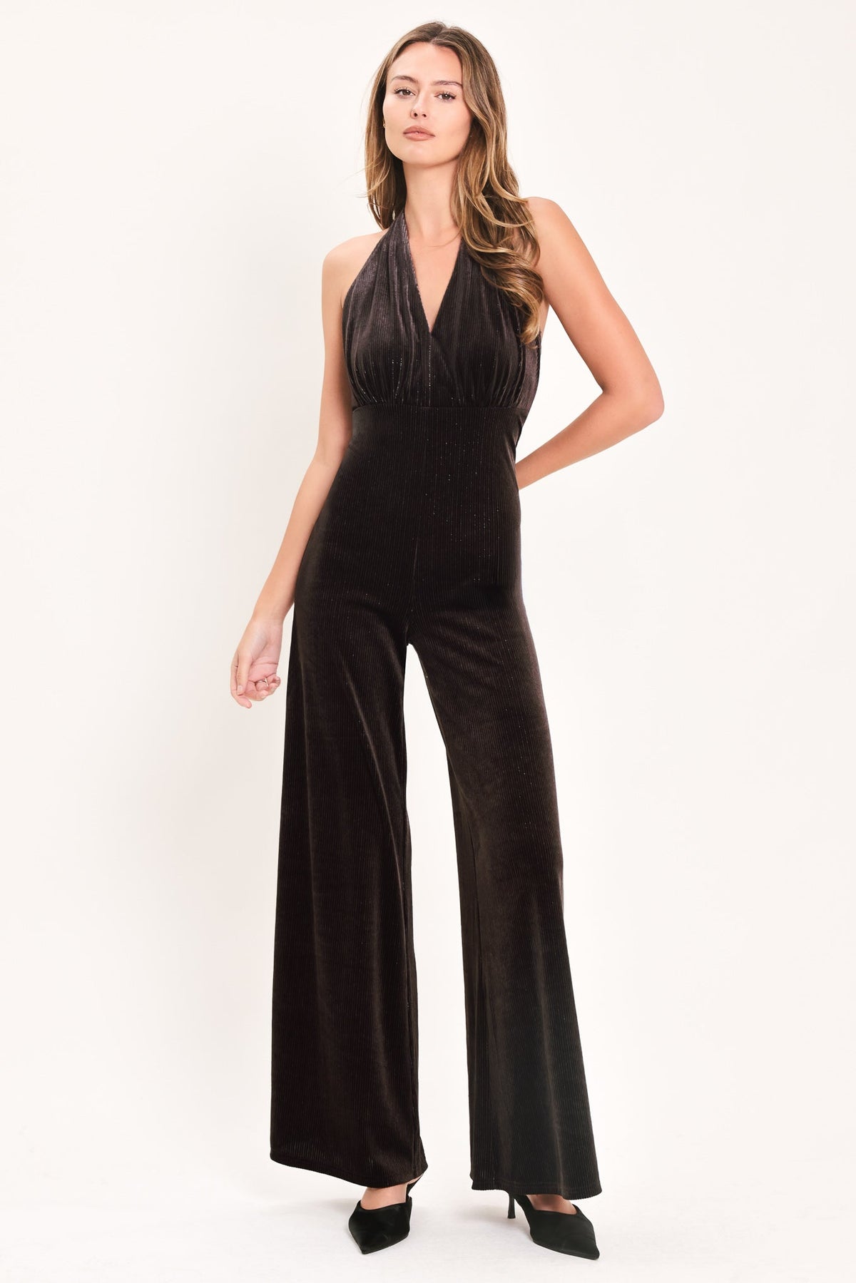 A woman modeling a black velvet halter jumpsuit with wide-leg pants, photographed against a plain white studio background.