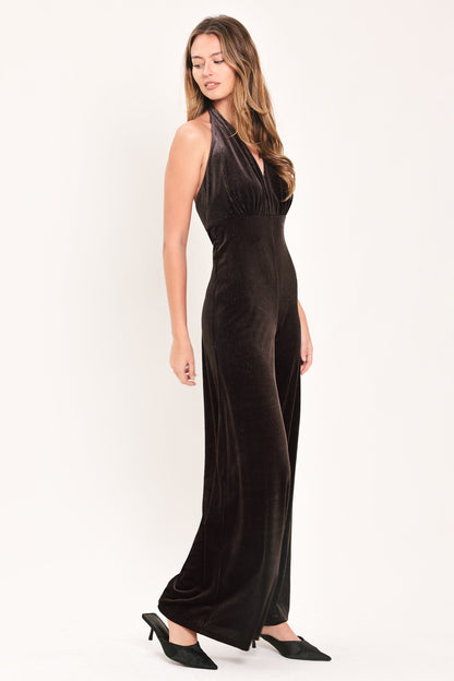 A woman modeling a sleeveless, floor-length black velvet halter gown with a deep V-neckline and draped side details, paired with black pointed-toe flats, against a plain white studio background.