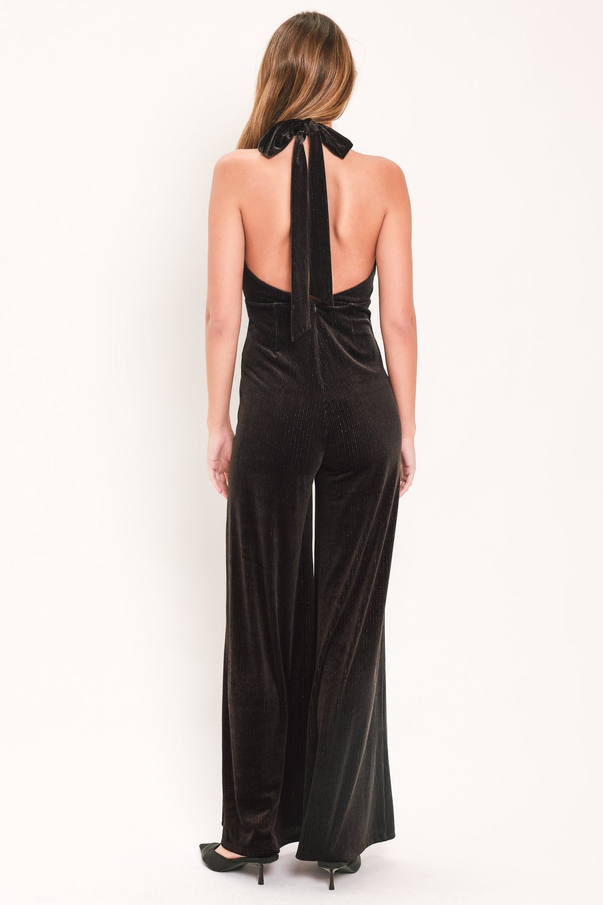 A woman viewed from the back wearing a black velvet halter jumpsuit with wide legs and a bow detail at the neck, set against a plain white studio background.