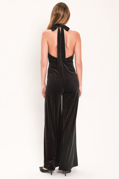A woman viewed from the back wearing a black velvet halter jumpsuit with wide legs and a bow detail at the neck, set against a plain white studio background.