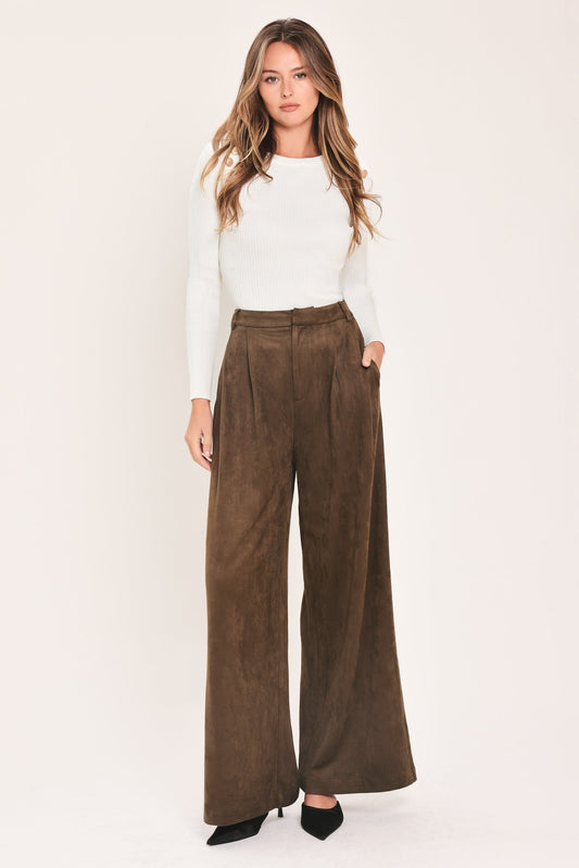 A woman modeling a pair of high-waisted, wide-leg brown suede pants with a white long-sleeve top and black heels, standing against a plain off-white studio background.