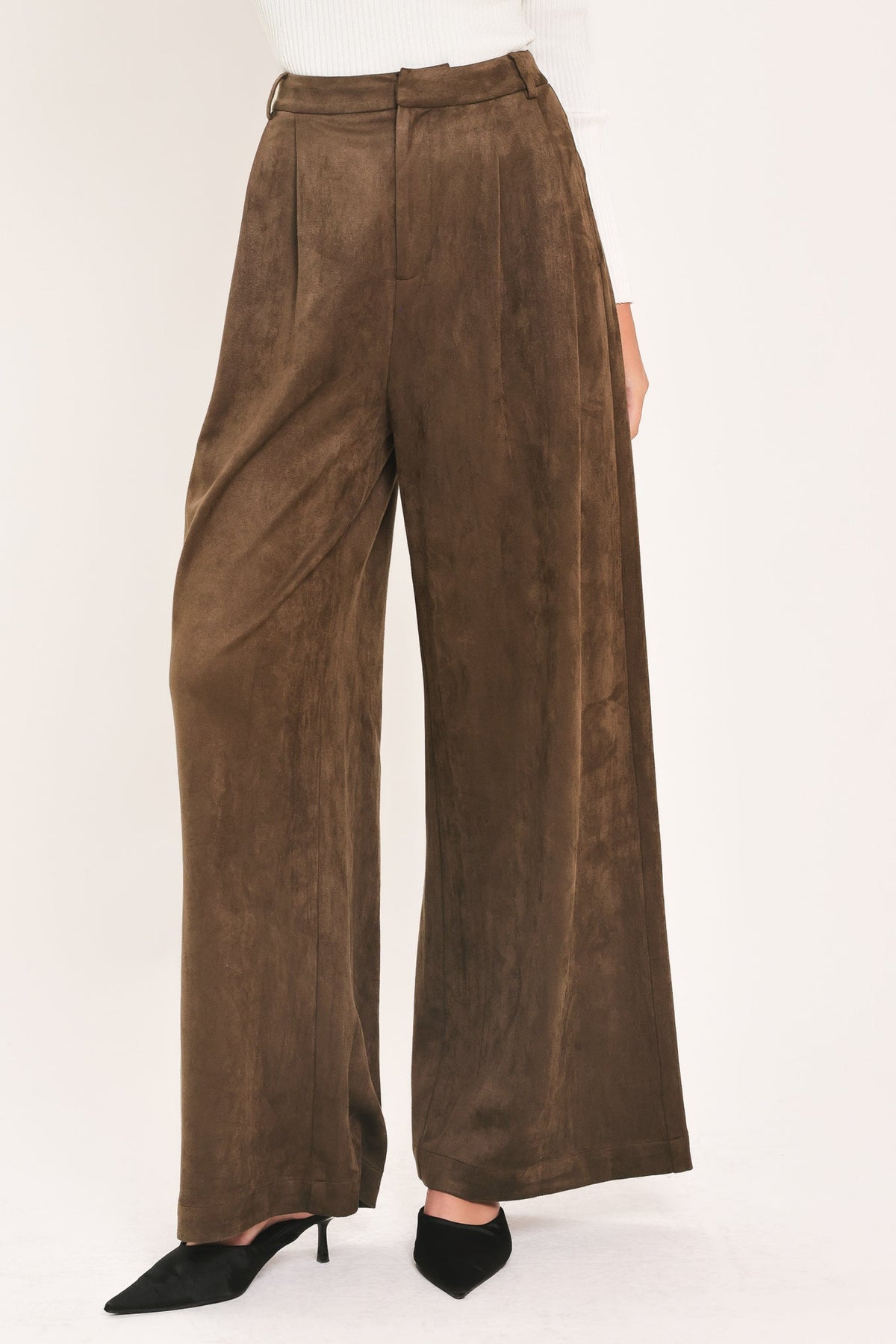 A person wearing high-waisted, wide-leg brown suede pants with black pointed-toe heels, shown against a plain light background.
