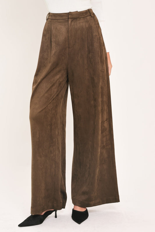 A person wearing high-waisted, wide-leg brown suede pants with black pointed-toe heels, shown against a plain light background.