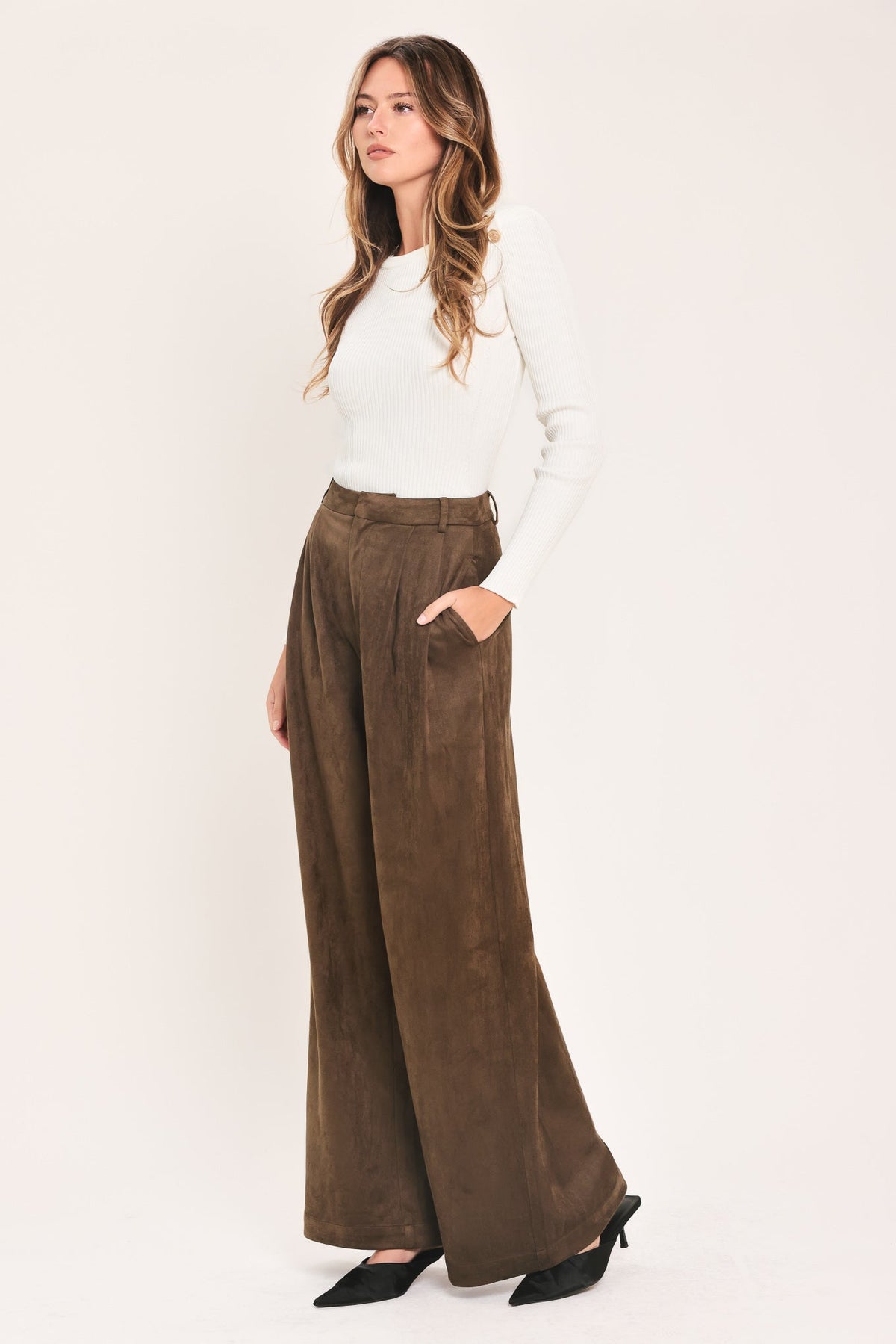 A woman modeling a pair of wide-leg brown corduroy pants with a white long-sleeve top and black pointed-toe heels, standing against a plain off-white studio background.